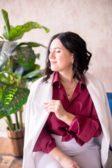 A young girl stylist with dark hair smiles in a trendy white jacket, a burgundy color shirt and light trousers in a light studio with green living plants in pots
