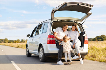 Image of happy mother and father traveling together with their infant daughter by comfortable car, having rest drinking tea or coffee from thermos, enjoying vacation.