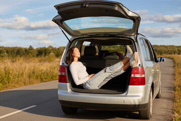 Outdoor shot of relaxed smiling pretty woman wearing white shirt and jeans sitting in car trunk on sunny summer day closed eyes, resting after long hour steering.