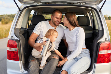 Outdoor shot of cheerful family traveling together by car, drinking tea or coffee from thermos...