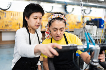 Two women checking a bicycle in a garage, a woman in her forties and an Asian girl in her twenties,...