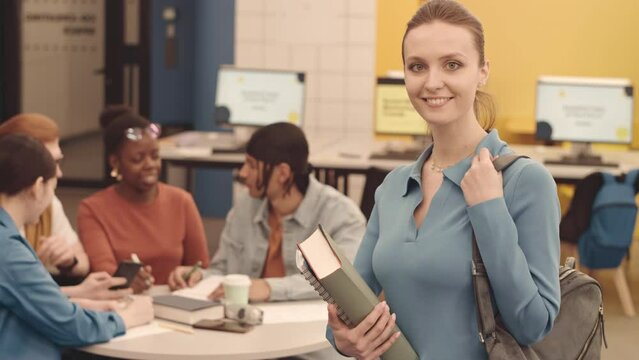 Tilt up medium slowmo portrait of Caucasian female student smiling at camera standing in modern coworking space at university with her multicultural peers chatting at table in background