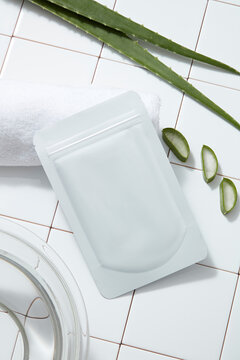 A facial sheet mask packaging mockup, white towel and fresh aloe vera leaves on white tile floor. Background for advertising cosmetic of aloe vera extract with bathroom concept.