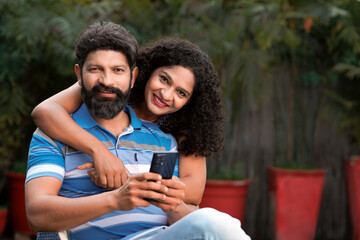 Happy indian couple sitting at out of home and using smartphone together.