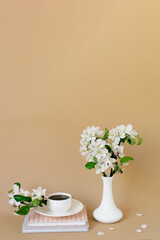 Romantic spring still life on a beige background. A vase with white apple blossoms, a cup of tea in a white porcelain cup and books