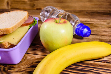 Ripe apple, banana, bottle of water and lunch box with sandwiches on wooden table