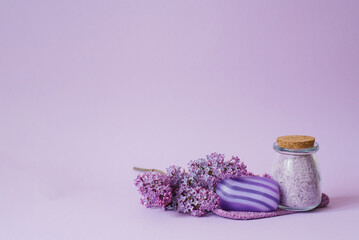 Cosmetic spa set for body care. Sea salt in glass jar, organic soap, washcloth glove and lilac flowers on purple background with copy space