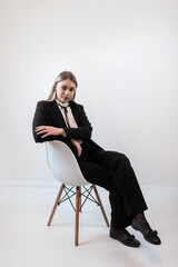 Fashion portrait of young blonde caucasian woman in trendy suit sitting on chair isolated on white