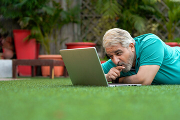 Indian senior man laying down at ground and using laptop.