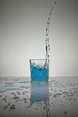 Vertical shot of water pouring into crystal glass with  droplets around, on a white shiny table