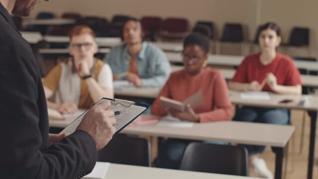 Cropped shot of Caucasian bearded university professor holding notes on clipboard while giving lecture to multicultural group of students sitting at desks in auditorium