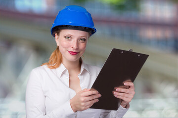 Young female engineer wearing a blue hard hat is working with a note pad in front of a construction site