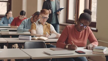 Waist up slowmo of group of multiethnic college students sitting one by one at desk in classroom writing test