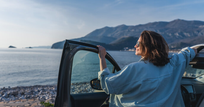 Young Happy Woman Traveler Enjoying The Sunset At The Sea While Standing Next To The Car. Summer Holidays And Travel Concept