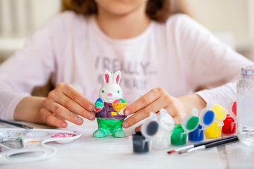  Girl at table showing colored statue of rabbit, children activity, drawing with with paints and a brush. No visible face.