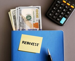 Blue notebook, pen calculator, cash money with handwritten text note REINVEST, to put money that...