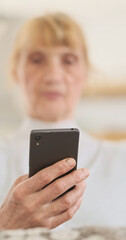 Elderly caucasian woman, using smartphone, holding it in front of her, close-up, vertical video, copy space. Concept of using new devices by old people, grandmothers, mothers.