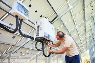 Young man adjusting hybrid inverter. Worker connecting wires to PV battery. Alternative energy in...