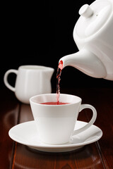 Red tea is poured from a white teapot into a white cup on a black background. Porcelain teaware. Side view. Selective focus.
