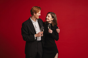 Smiling couple holding glasses with champagne while standing isolated over red wall