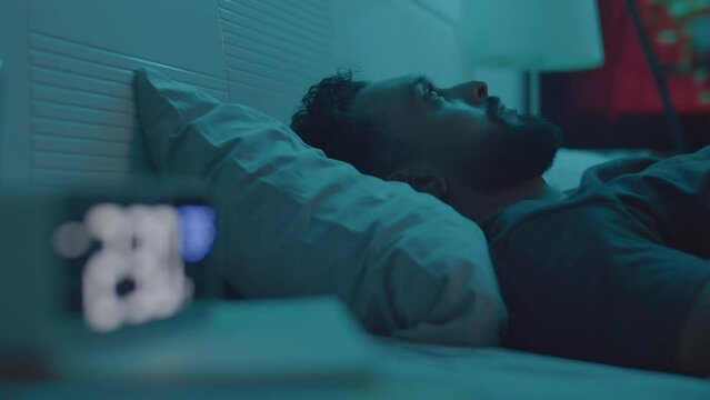 Rack focus shot of young man lying in bed in dark room and looking at digital clock while suffering from insomnia during night