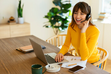 Happy freelancer talking on headset sitting with laptop at home office
