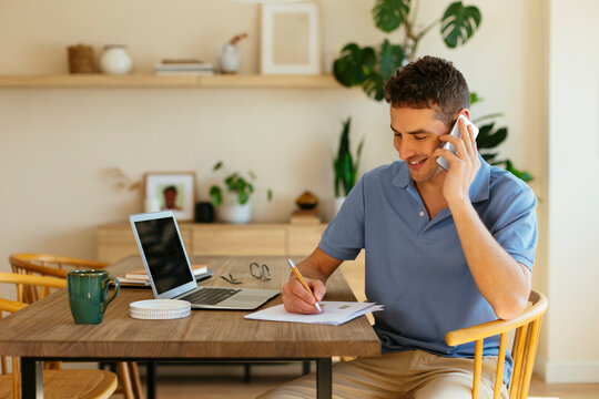 Smiling freelancer talking on mobile phone and taking notes on paper at table