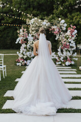 Fototapeta na wymiar Bride in a white long dress walking to a fresh flowers decorated arch against the background of trees. An elegant outdoor country ceremony.