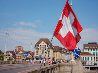 Naklejka premium Beautiful Swiss flag is waving front of historical street in Basel, Switzerland, Johanniter Bridge