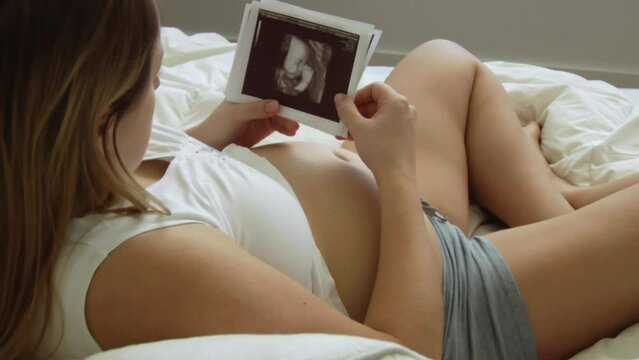 Pregnant woman sitting on the bed and looking at ultrasound pictures of her baby