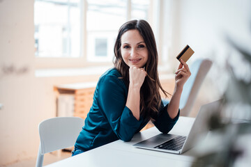 Smiling businesswoman holding credit card sitting at table in home office