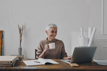 Happy senior businesswoman enjoying hot drink while using laptop at her working place