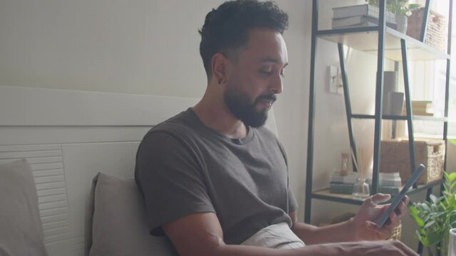 Young bearded man having breakfast in bed, scrolling through social media on smartphone and smiling in morning at home