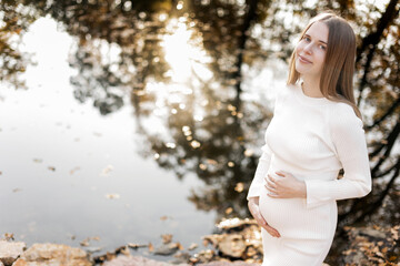 Mothers day. Beautiful young pregnant woman in white tight knit dress with little belly relaxing outdoors by the pond with reflection nature and sun in the park and looking at camera. Women day