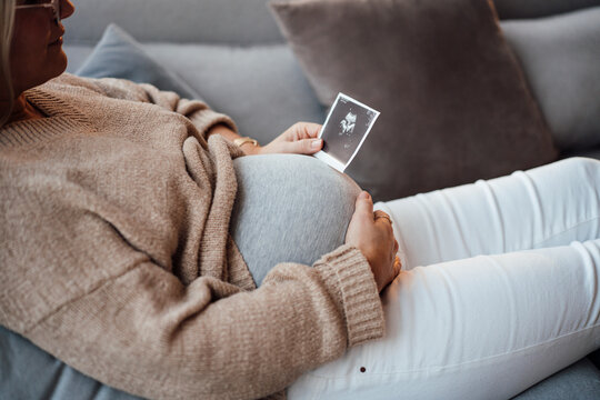 Pregnant woman holding ultrasound image sitting on sofa at home