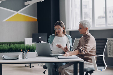 Two confident women using technologies and communicating while working in office together