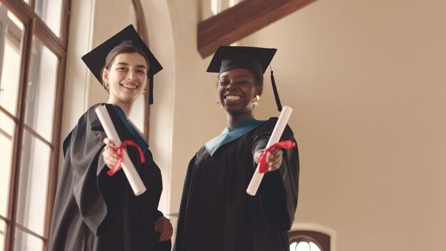 Low angle slowmo portrait of two happy diverse student girls in graduation gowns showing their diplomas with red ribbons at camera and smiling