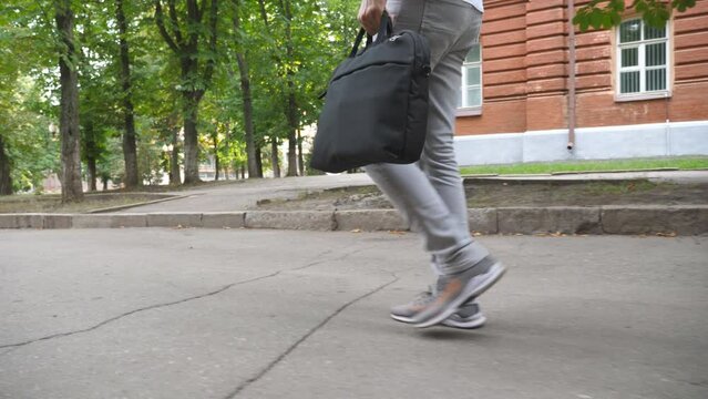Legs of young man in casual wear going through urban park. Businessman with bag walking at urban street. Guy on his way study or to meeting in office. Concept of morning commute. Slow motion Close up