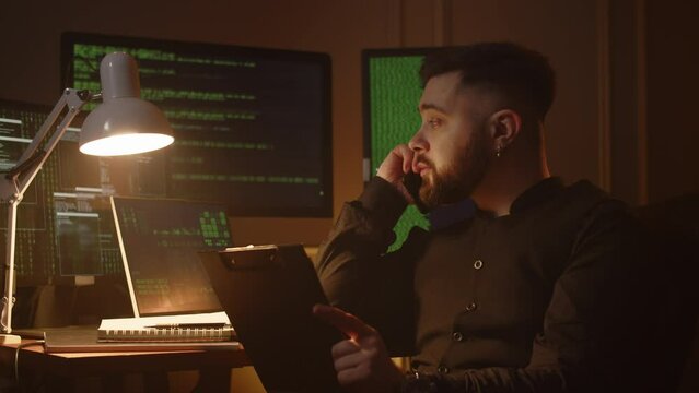 Video of a man holding a clipboard and talking on the phone. A programmer is discussing the details of developing a new application with his colleagues over the phone.