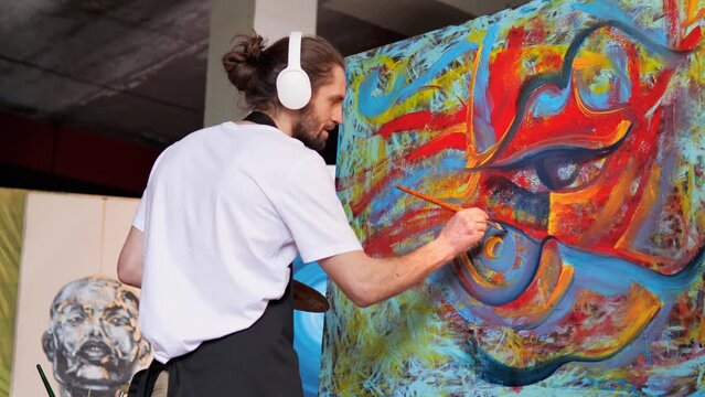 Focused artist with a creative, innovative spirit, adorned with long hair, headphones, black apron stained with paint, crafting magnificent masterpiece in art studio. Colors of red, blue, and yellow.