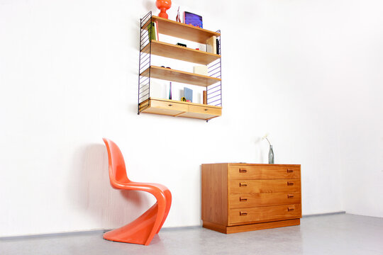 a mid century String Shelf with drawers original from the 60s mounted on a white wall with books and other accessories with a commode made of teak wood scandinavian design orange chair living room 60s
