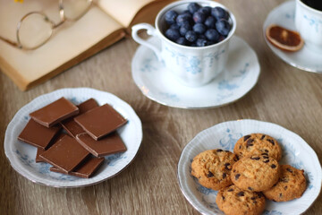 Cup of tea or coffee, plate of cookies, cup of blueberries, plate of chocolate, glass of juice, book, reading glasses, teapot, flowers and lit candles on the table. Brunch or afternoon tea concept.