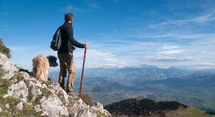 Hombre joven barbudo  con palo de madera y perro en montaña de Asturias mirando el horizonte
