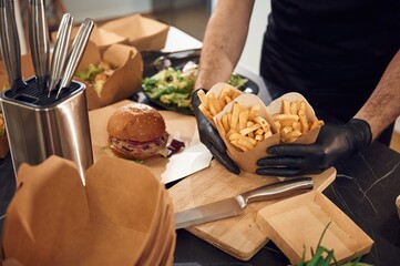 French fries, vegetables and burger. Man is packing food into the paper eco boxes. Indoors, restaurant