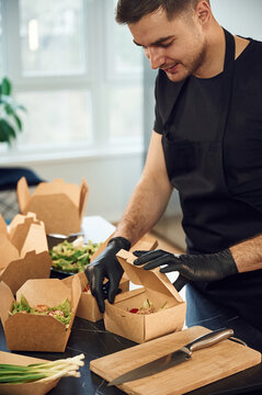 View from the side of meal, vegetables. Man is packing food into the paper eco boxes. Indoors, restaurant