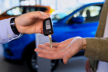 Close-up of the hands of the seller who is handing over the car key to the buyer against the background of cars in the showroom