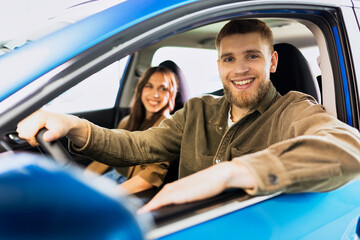 A happy man is driving a car at a dealership. A young woman is sitting in the passenger seat