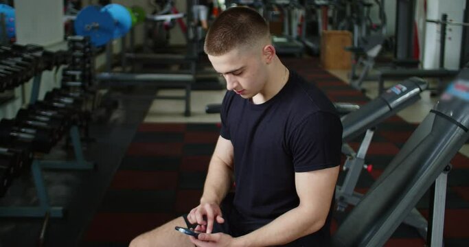 Portrait of guy in gym while resting between a workout uses phone body strength fit health male strong healthy muscular athlete guy physical young arm model mobile caucasian endurance close up.