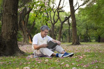 Fototapeta premium old man with grey hair wears glasses sitting under a tree and reading a book in forest park, mature male spend his free time in nature, concept of elderly lifestyle in nature,hobbies,relaxing,resting