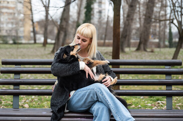 Young beautiful hipster woman sitting on a bench in the park with her adopted dog that saved his life from the street and the animal shelter and keeps him in her arms, gives him love food and care